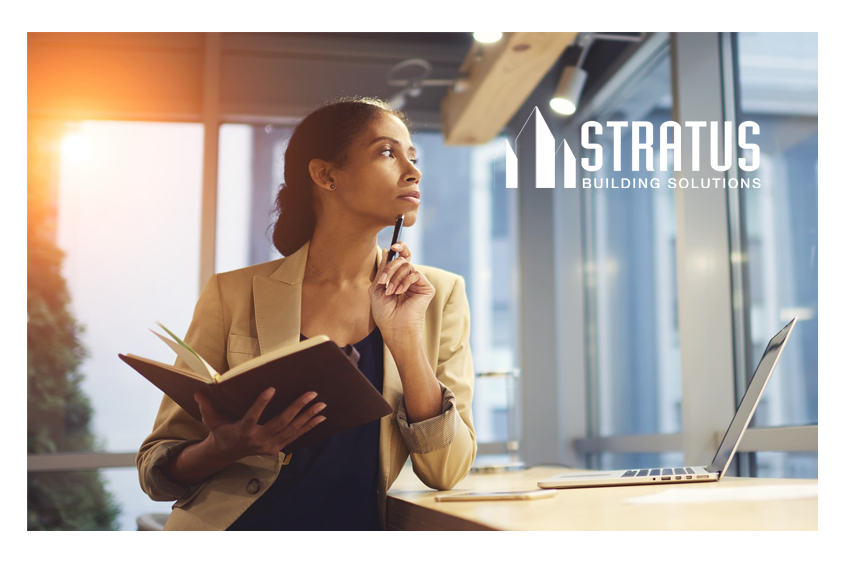 A woman in business attire stands next to a desk thoughtfully holding a pen up to her chin while holding a notebook 
