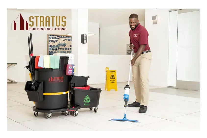A Smiling Uniformed Stratus Cleaner with a Trash Can and Mop Bucket on Wheels with Cloths on it Next to Him Mops a Tile Floor