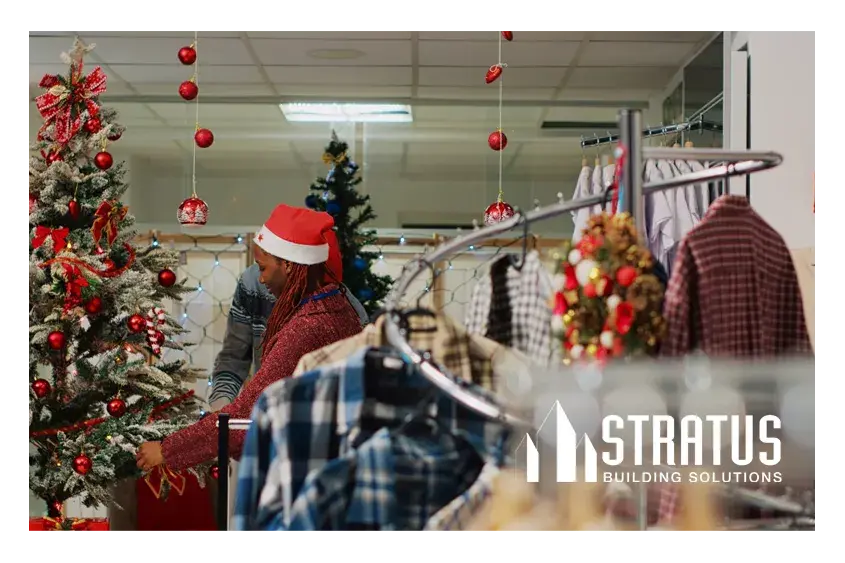 A rack of clothes in front a Christmas tree and a retail worker decorating a store for Christmas. 